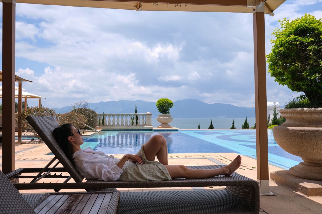 about-02 A woman in sunglasses relaxes on a sunlounger by a luxury poolside with a scenic view.
