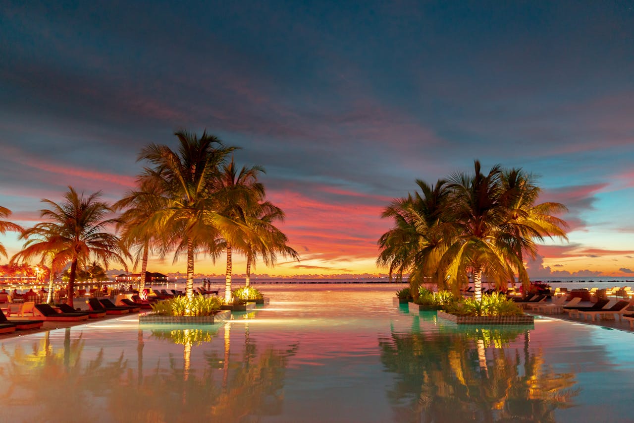 why-choose-us Beautiful sunset over a tropical resort pool with palm trees and vibrant sky reflections.